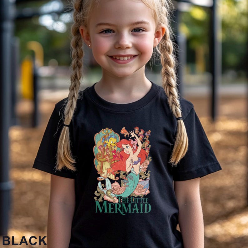 May include: Black t-shirt featuring a colorful graphic of Ariel from The Little Mermaid with the text "THE LITTLE MERMAID" in green. The shirt is worn by a young girl with blonde braids.