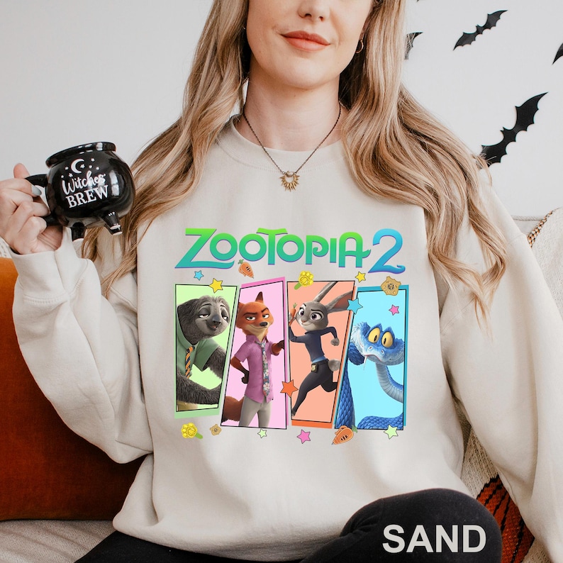 Może przedstawiać: Beżowa bluza z napisem "ZOOTOPIA 2" w kolorze zielonym i niebieskim. Projekt zawiera kresk&oacute;wkowe obrazy postaci z filmu. Słowo "SAND" jest wydrukowane na dole bluzy.