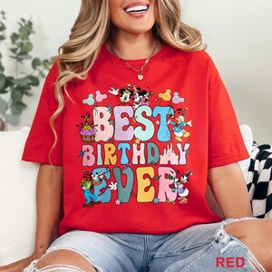 May include: Red t-shirt with the text "BEST BIRTHDAY EVER" in colorful bubble letters. The design includes cartoon characters like Mickey and Minnie Mouse, Donald Duck, and Goofy, with a birthday cake and confetti.