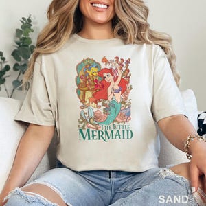 May include: Sand-colored t-shirt featuring a colorful graphic of Ariel from The Little Mermaid. The graphic includes Ariel, Flounder, and floral elements. The text "THE LITTLE MERMAID" is printed in green below the graphic. The word "SAND" is printed in the bottom right corner.