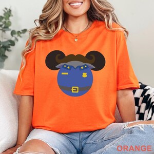 Puede incluir: Camiseta naranja con un dise&ntilde;o de dibujos animados de la cabeza de un polic&iacute;a azul con bigote y orejas negras. La camiseta tiene la palabra "ORANGE" impresa en la parte inferior. La camiseta es de material suave.