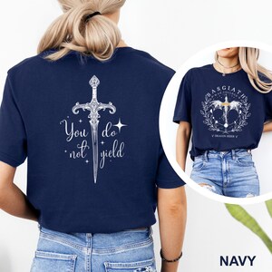May include: Navy blue t-shirt featuring a detailed white sword design with the words "You do not yield." A second image shows a similar shirt with a circular design and the text "Basgiath War College Dragon Rider."
