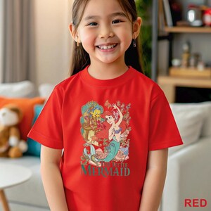 May include: A red t-shirt featuring a graphic of Ariel and Flounder from The Little Mermaid. The design includes the text "THE LITTLE MERMAID" in green. The shirt is being worn by a young girl.