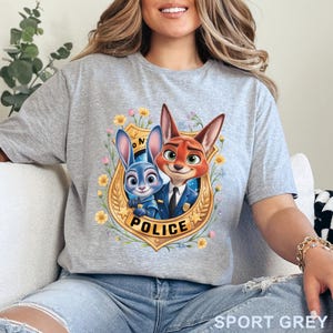 Peut inclure: T-shirt gris sport avec un motif de dessin anim&eacute; repr&eacute;sentant un lapin bleu et un renard roux &agrave; l'int&eacute;rieur d'un badge de police dor&eacute;. Le badge est entour&eacute; de fleurs jaunes et de feuilles vertes. Le mot "POLICE" est imprim&eacute; sur le badge.