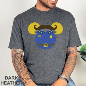 Puede incluir: Camiseta gris jaspeado oscuro con un dise&ntilde;o de polic&iacute;a de dibujos animados. El dise&ntilde;o incluye una placa azul, un bigote marr&oacute;n y cuernos de carnero amarillos. El texto "DARK HEATHER" est&aacute; impreso en la parte inferior de la camiseta.