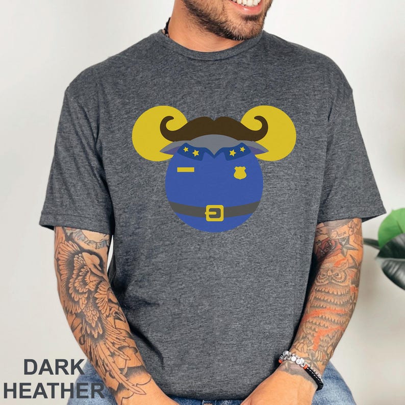 Puede incluir: Camiseta gris jaspeado oscuro con un dise&ntilde;o de polic&iacute;a de dibujos animados. El dise&ntilde;o incluye una placa azul, un bigote marr&oacute;n y cuernos de carnero amarillos. El texto "DARK HEATHER" est&aacute; impreso en la parte inferior de la camiseta.