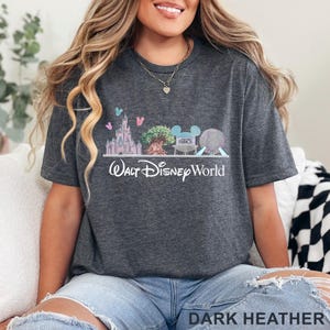 May include: Dark heather gray t-shirt featuring a Walt Disney World graphic. The design includes a castle, tree, Mickey Mouse ears, and a sphere. The words "Walt Disney World" are printed below the graphic.