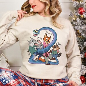 K&ouml;nnte beinhalten: Sandfarbenes Sweatshirt mit einer Cartoon-Grafik von Zootopia-Charakteren, darunter Nick, Judy und andere, die sich um eine blaue Zahl 2 wickeln. Das Sweatshirt hat lange &Auml;rmel und einen Rundhalsausschnitt. Das Wort "SAND" ist unten aufgedruckt.