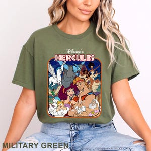 May include: Military green t-shirt featuring a colorful graphic from Disney's Hercules. The design includes characters like Hercules, Megara, Pegasus, and Hades. The shirt has a classic crew neck and short sleeves.