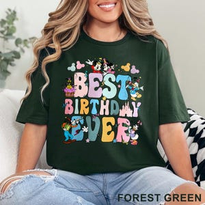 May include: Forest green t-shirt with the text "BEST BIRTHDAY EVER" in colorful bubble letters. The design includes cartoon characters like Mickey and Minnie Mouse, Donald Duck, and Goofy. The shirt is a relaxed fit.