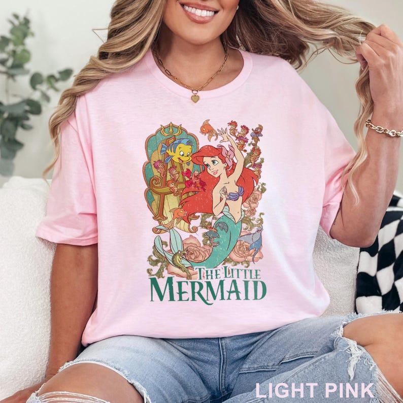 May include: Light pink t-shirt featuring a colorful graphic of Ariel and Flounder from The Little Mermaid. The graphic includes the text "The Little Mermaid" in green. The shirt is made of a soft material.