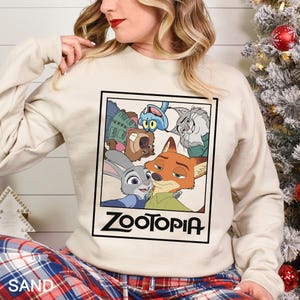 Peut inclure: Un sweat-shirt couleur sable avec une image encadr&eacute;e des personnages du film d'animation "Zootopie". L'image comprend Judy Hopps, Nick Wilde et d'autres personnages. Le mot "ZOOTOPIA" est imprim&eacute; en gras, en lettres noires sous l'image.