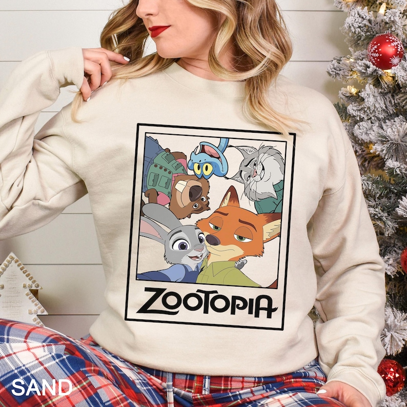 Peut inclure: Un sweat-shirt couleur sable avec une image encadr&eacute;e des personnages du film d'animation "Zootopie". L'image comprend Judy Hopps, Nick Wilde et d'autres personnages. Le mot "ZOOTOPIA" est imprim&eacute; en gras, en lettres noires sous l'image.