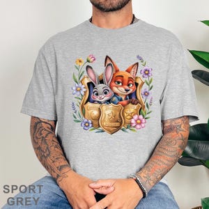 Puede incluir: Camiseta gris deportiva con un dise&ntilde;o colorido de Judy Hopps y Nick Wilde de Zootopia, enmarcado por flores y placas de polic&iacute;a. El dise&ntilde;o incluye el texto "Zootopia Police".