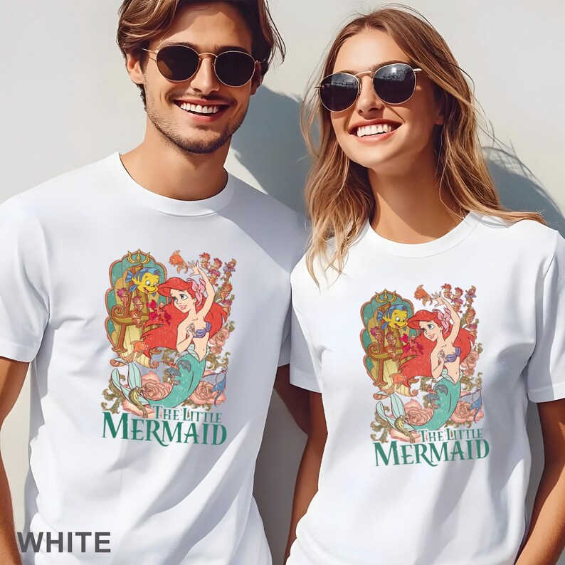 May include: White t-shirts featuring a colorful graphic of Ariel from The Little Mermaid, with the text "THE LITTLE MERMAID" in green. The design includes Ariel, Flounder, and Sebastian, surrounded by floral elements. The word "WHITE" is at the bottom.