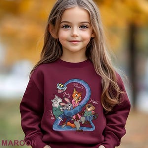 K&ouml;nnte beinhalten: Burgunderfarbenes Sweatshirt mit einem Cartoon-Motiv von Zootopia-Charakteren, darunter Judy Hopps und Nick Wilde, und ihren Namen. Das Wort "MAROON" ist unten aufgedruckt. Das Sweatshirt wird von einem jungen M&auml;dchen getragen.