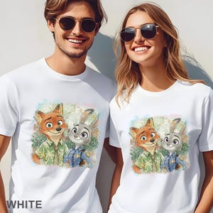 K&ouml;nnte beinhalten: Wei&szlig;e T-Shirts mit einer farbenfrohen Illustration von Nick Wilde und Judy Hopps aus Zoomania. Die Zeichentrickfiguren sind vor einem Hintergrund im Aquarellstil platziert. Das Wort "WHITE" ist unten im Bild aufgedruckt.