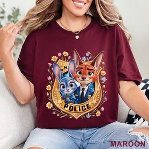 Peut inclure: T-shirt bordeaux avec une illustration de dessin anim&eacute; d'un lapin bleu et d'un renard roux &agrave; l'int&eacute;rieur d'un &eacute;cusson de police dor&eacute;. L'&eacute;cusson est entour&eacute; de fleurs jaunes et bleues, avec le mot "POLICE" en bas.