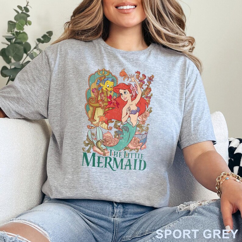 May include: Sport grey t-shirt featuring a colorful graphic of Ariel from The Little Mermaid, with the text "THE LITTLE MERMAID" in green. The design includes floral elements and characters from the movie.