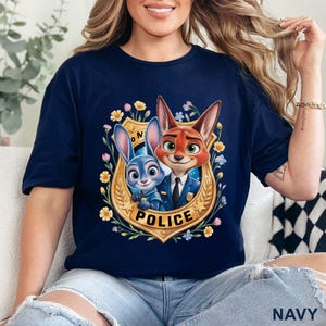 Peut inclure: T-shirt bleu marine avec un motif color&eacute; de Judy Hopps et Nick Wilde de Zootopie en uniforme de police. Le dessin est encadr&eacute; par un &eacute;cusson dor&eacute; avec le mot "POLICE" et des fleurs. Le t-shirt est en tissu doux.