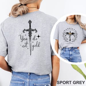 May include: Sport grey t-shirt featuring a detailed black sword design with the text "You do not yield" on the back. The front of the shirt displays a circular graphic with text and a sword design. The shirt is made of a soft material.