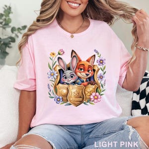 Puede incluir: Camiseta rosa claro con un dise&ntilde;o colorido de Judy Hopps y Nick Wilde de Zootopia, enmarcado por flores y una placa de polic&iacute;a. El texto "LIGHT PINK" est&aacute; en la parte inferior de la camiseta.
