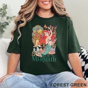 May include: Forest green t-shirt featuring a colorful graphic of Ariel and Flounder from The Little Mermaid. The design includes floral elements and the movie title. The shirt is a relaxed fit.