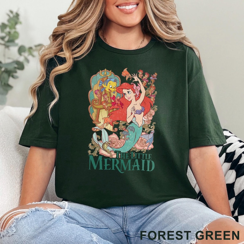 May include: Forest green t-shirt featuring a colorful graphic of Ariel and Flounder from The Little Mermaid. The design includes floral elements and the movie title. The shirt is a relaxed fit.