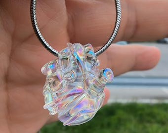 Puede incluir: Un colgante de cristal transparente e iridiscente con forma de corazón, suspendido de una cadena plateada. El colgante refleja un arcoíris de colores. La cadena es de diseño fino tipo serpiente.