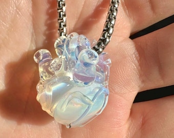 Puede incluir: Un colgante de vidrio transparente e iridiscente en forma de corazón, suspendido de una cadena plateada. El colgante presenta un diseño texturizado y orgánico con detalles arremolinados y un sutil juego de colores.