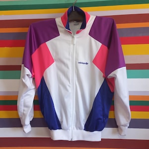 May include: A vintage Adidas track jacket in white, purple, pink, and blue. The jacket has a full zipper, a stand-up collar, and the Adidas logo embroidered on the chest. The jacket is hanging against a colorful striped background.