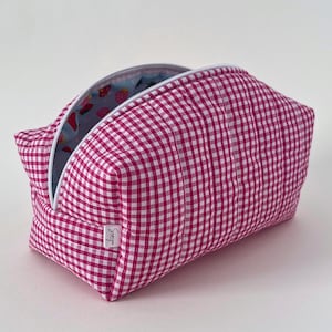 May include: A pink and white gingham patterned cosmetic bag with a white zip. The interior lining is light blue with a strawberry pattern. A small white tag with the word "Savvy" is sewn on the side.