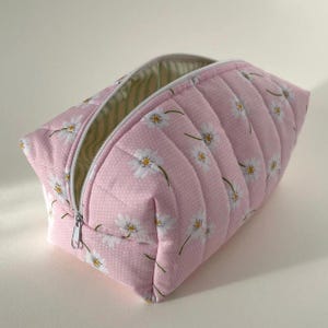 May include: A pink quilted cosmetic bag with a white daisy floral pattern and a white zip. The interior lining is striped in green and white. The bag is open, showing the inside.