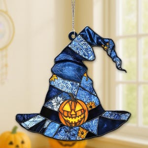 May include: A blue witch hat ornament with a jack-o'-lantern design. The hat features a patchwork pattern with floral and swirl details. The pumpkin has a carved face and is illuminated with an orange glow. A silver chain is attached for hanging.