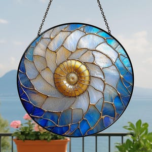 May include: Round stained glass window with a nautilus shell design. The window features a spiral pattern in shades of blue, white, and gold, with a black metal frame and chain for hanging. The background shows a lake and mountains.