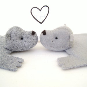 May include: Two grey felt animal figures facing each other, each featuring small black bead eyes and a dark brown stitched nose. Above the figures is a hand-drawn black outline of a heart shape on a white background with a subtle grid pattern.