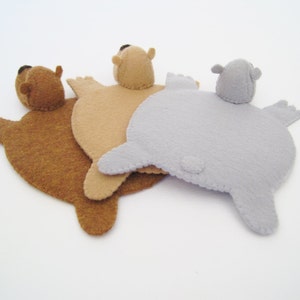 Pode incluir: Três porta-copos ou ornamentos planos de feltro em forma de animal nas cores castanho, bege e cinzento-claro. Cada peça apresenta um corpo arredondado, quatro patas pequenas, uma cauda pequena e uma cabeça com duas orelhas e um nariz escuro.