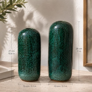 Könnte beinhalten: Zwei grüne Keramikvasen. Die größere Vase ist 28 cm hoch, und die kleinere Vase ist 27 cm hoch. Beide Vasen haben eine zylindrische Form mit einer strukturierten, glänzenden Oberfläche. Der Sockel jeder Vase ist 13 cm breit.
