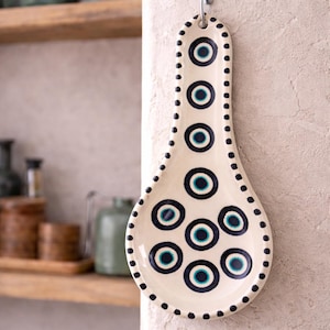 May include: Cream-colored ceramic spoon rest with a pattern of navy blue and teal circles. The spoon rest is hanging on a wall. The edges are dotted with small navy blue circles. A decorative kitchen item.