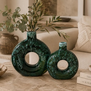 Pode incluir: Dois vasos de cerâmica verde escuro com uma abertura circular, um grande e um pequeno, são exibidos em uma superfície de cor clara. Os vasos têm um padrão salpicado e são preenchidos com vegetação. Uma tigela de limas e livros estão por perto.