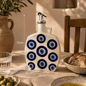 Puede incluir: Una botella dispensadora de aceite de cerámica blanca con un patrón repetitivo de ojos azules y blancos. La botella tiene un vertedor de color plateado y un asa. Mide 25 cm de alto y 16 cm de ancho, según las marcas visibles.