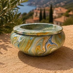 Pode incluir: Uma taça redonda de perfil baixo com tampa, apresentando um padrão marmoreado com redemoinhos de cores verde, azul, branco e amarelo ocre. O objeto tem um acabamento liso e brilhante e uma borda larga e plana na tampa.