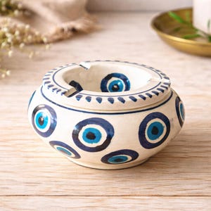 May include: A cream-coloured ceramic ashtray decorated with blue eye motifs. The rim is adorned with small blue dashes. The circular ashtray has a recessed area for holding ashes and cigarettes, and is a decorative item.