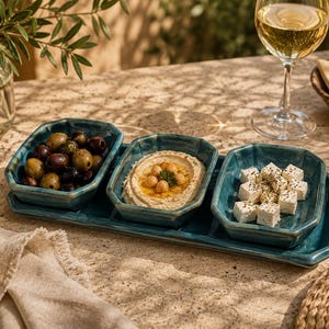 Puede incluir: Una bandeja de servicio rectangular de cerámica color verde azulado con tres cuencos octogonales integrados. Los cuencos contienen aceitunas oscuras y verdes, hummus cremoso con garbanzos y cubos de queso feta blanco con hierbas.
