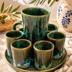 May include: A ceramic drink set featuring a tall pitcher and five matching tumblers arranged on a round tray. The pieces have a reactive glaze finish in deep forest green, teal, and dark brown tones with a mottled, dripped texture throughout.