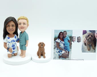 Puede incluir: Una colección de regalos personalizados, que incluye figuras de bobblehead de una pareja y un perro, junto con un collage de fotos enmarcado. Las figuras están sobre bases blancas, y la foto muestra a una familia en la playa.