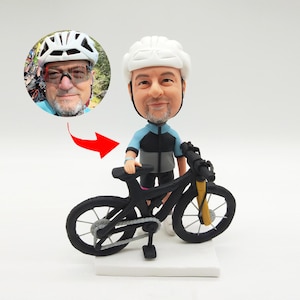 May include: A custom figurine of a person wearing a white helmet and blue and grey cycling jersey, standing next to a black mountain bike. The figurine is holding the handlebars of the bike.