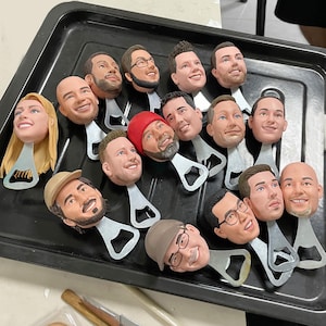 May include: A selection of custom bottle openers, each with a sculpted head depicting different people. The heads are attached to a silver bottle opener. Some heads wear hats or glasses. The openers are arranged on a black tray, ready for use.
