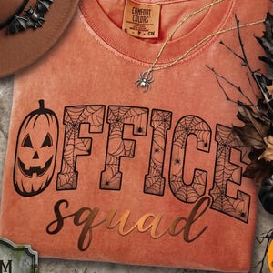 May include: A burnt orange t-shirt featuring the text OFFICE SQUAD in black, with the letter O replaced by a jack-o'-lantern and the other letters filled with spiderweb patterns. A silver spider necklace rests on the neckline.