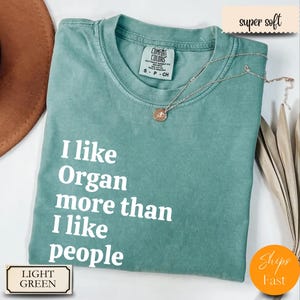 May include: Light green Comfort Colors t-shirt with the text "I like Organ more than I like people" in white. A silver necklace with a pendant is on the shirt. The shirt is labeled "super soft".
